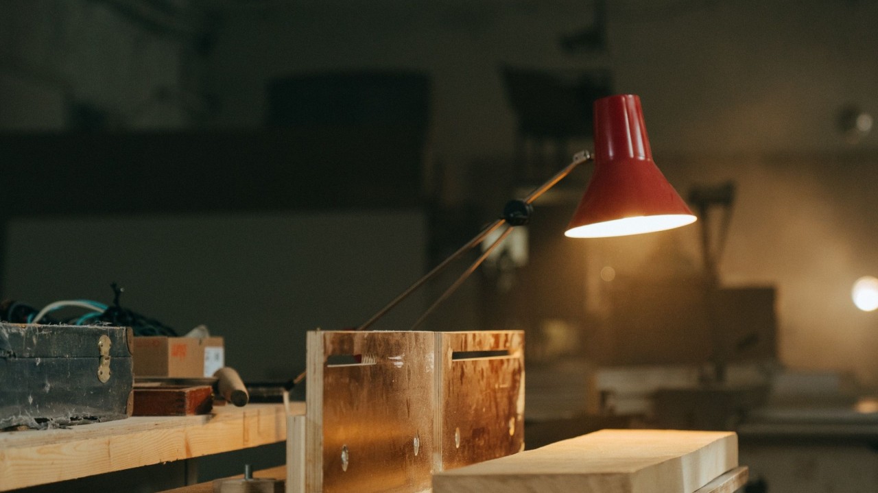 A red anglepoise desk lamp lighting a half built wooden box on a bench in a dim woodwork shop.